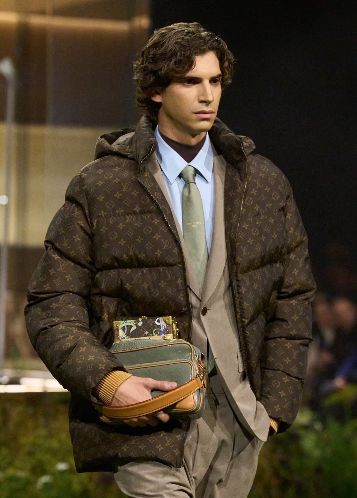 A male model walks down a runway wearing a brown Louis Vuitton monogram puffer jacket over a light blue dress shirt, olive tie, and beige suit jacket and trousers. He is holding a Louis Vuitton monogram shoulder bag.